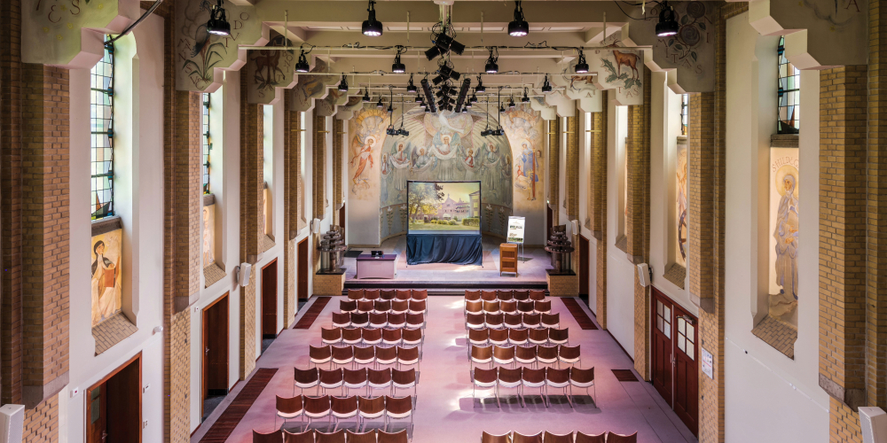 De Ceciliakapel in het klooster van Conferentiehotel Kontakt der Kontinenten, een voormalige kapel met hoge wanden, originele muurschilderingen en glas-in-loodramen, ingericht in theateropstelling met rijen stoelen gericht op het podium.