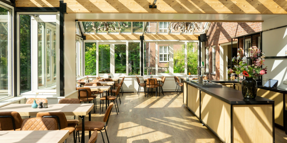 De Wintertuin van Conferentiehotel Kontakt der Kontinenten, een lichte ruimte met veel glas, houten plafondelementen, tafels en stoelen, en uitzicht op het groen rondom het klooster.
