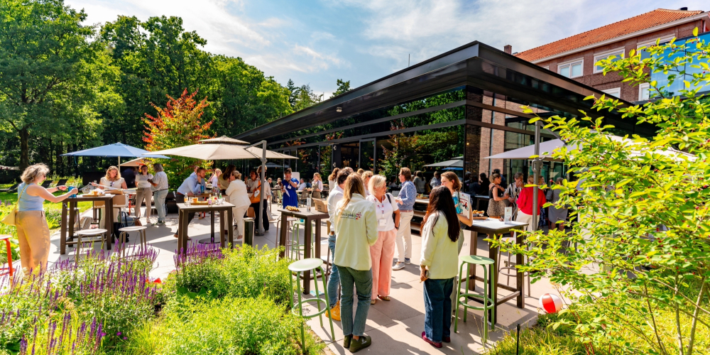 Het terras van restaurant KonneKt bij Conferentiehotel Kontakt der Kontinenten, waar mensen staand met elkaar in gesprek zijn aan hoge tafels, omgeven door groen, parasols en het moderne restaurantgebouw in een bosrijke omgeving.