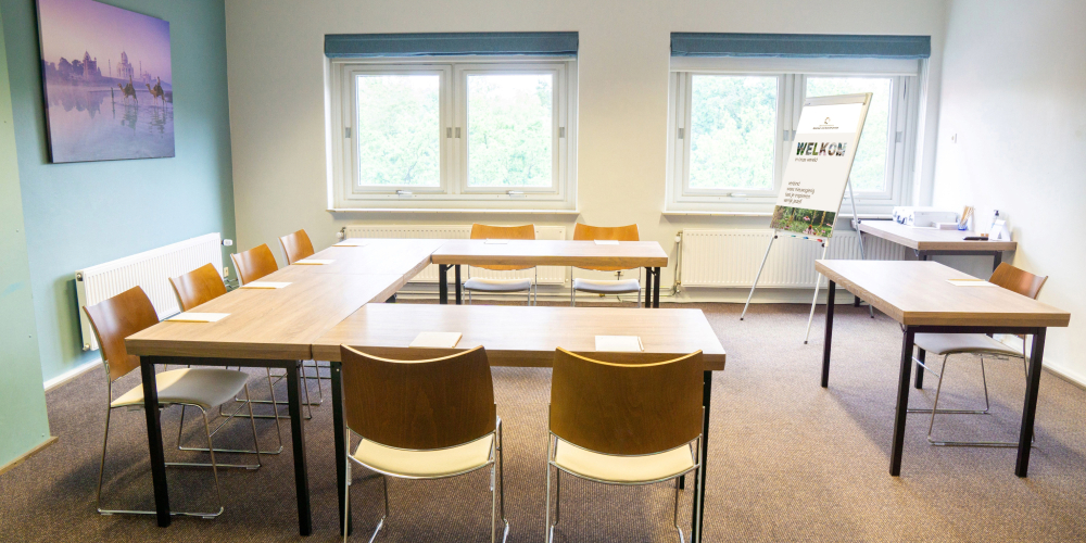 De vergaderzaal India van Conferentiehotel Kontakt der Kontinenten, met daglicht via twee ramen, houten tafels en stoelen opgesteld in een U-vorm, en een flip-over in de ruimte.