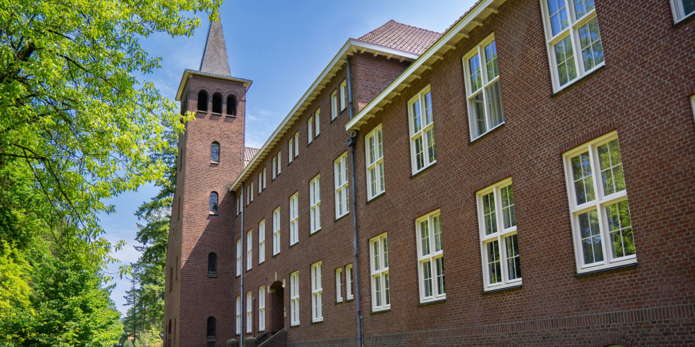 Klooster Cenakel van Conferentiehotel Kontakt der Kontinenten, een karakteristiek bakstenen gebouw met toren en hoge ramen, gelegen in een groene, bosrijke omgeving.