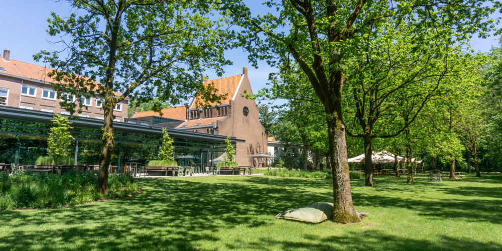 De bostuin en het terras van Conferentiehotel Kontakt der Kontinenten, met tafels en stoelen bij restaurant KonneKt, een zitzak op het gras op de voorgrond en een evenemententent op de achtergrond, omgeven door bomen in een bosrijke omgeving.