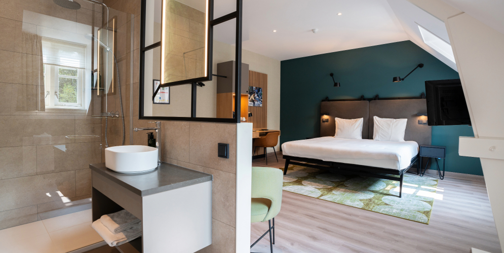 Een moderne tweepersoonskamer in het missiehuis van Conferentiehotel Kontakt der Kontinenten, met een comfortabel tweepersoonsbed, bureau, televisie en een open badkamer met douche en wastafel, afgewerkt in rustige kleuren en met veel daglicht.