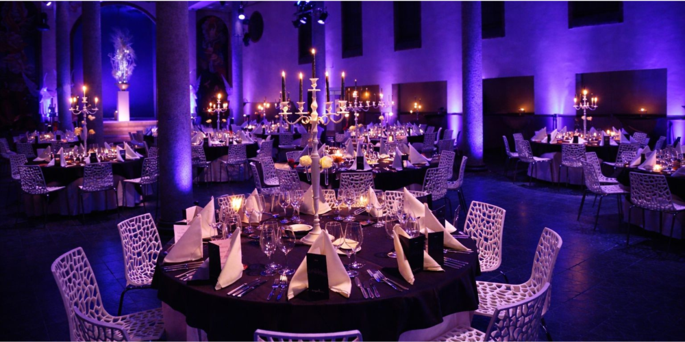 De foto toont een grote evenementenruimte in een kapel met een hoog, gebogen plafond dat rijk is beschilderd met klassieke en artistieke motieven. De ruimte is uitgelicht in blauwe tinten en voorzien van professionele verlichting en geluid. De diner opstelling en het hoge plafond geven een indrukwekkende, chique sfeer, geschikt voor diners, borrels en grootschalige evenementen.