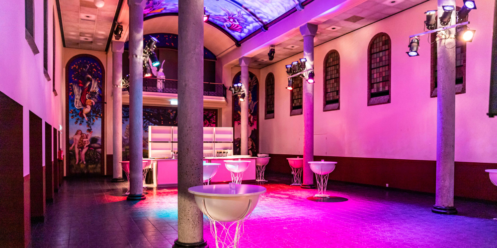 De foto toont een ruime evenementen locatie in een voormalige kapel met hoge zuilen, een beschilderd gewelfplafond en artistieke muurschilderingen. De zaal is verlicht met roze en paarse podiumverlichting. Er staan moderne, transparante statafels en achterin is een strak verlichte bar zichtbaar. De combinatie van historische architectuur en moderne inrichting maakt de locatie geschikt voor feesten, evenementen en culturele bijeenkomsten.