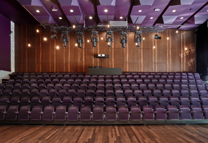 De foto toont de Eduard Flipse Zaal van de Doelen in Rotterdam. De zaal heeft een vaste tribuneopstelling met rijen paars gestoffeerde stoelen en een warme houten vloer. Achter het podium bevindt zich een houten wand met daarboven professionele theaterverlichting. Het plafond bestaat uit geometrische panelen in paarse tinten. De zaal biedt een intieme, moderne setting voor presentaties, lezingen en congressen.