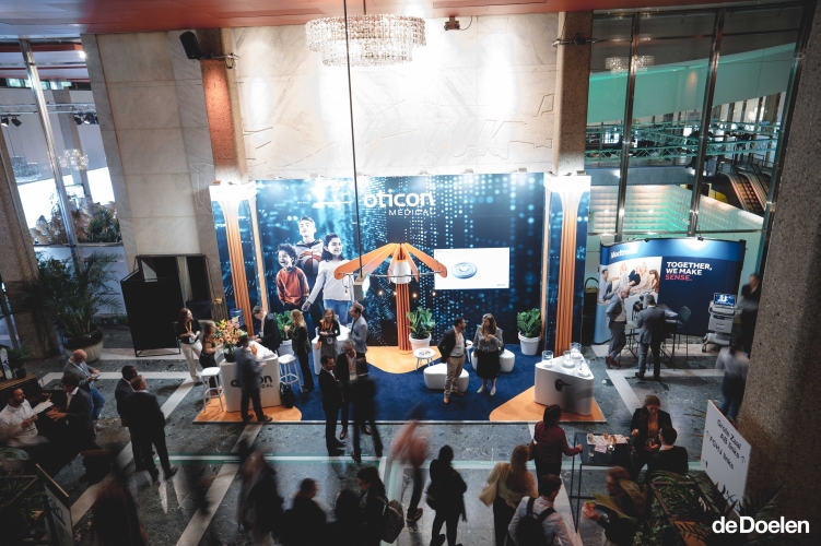 De foto toont de Grote Zaal Foyer van de Doelen in Rotterdam tijdens een congres. In de ruime foyer staan expositiestands en informatiebalies opgesteld, waar bezoekers met elkaar in gesprek zijn. De ruimte heeft een marmeren vloer, hoge kolommen, glazen wanden en een rood plafond met kroonluchters. De foyer is geschikt voor ontvangsten, exposities, netwerkmomenten en catering bij congressen en events.