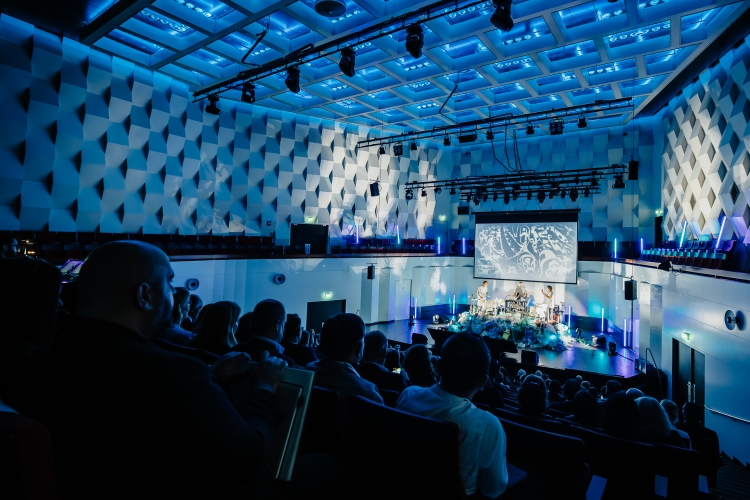 De foto toont de Jurriaanse Zaal van de Doelen in Rotterdam tijdens een congres of presentatie. Het publiek zit in vaste tribuneopstelling en kijkt uit op het podium waar sprekers staan onder blauwe en paarse podiumverlichting. De zaal heeft een karakteristieke architectuur met geometrische wand- en plafondpanelen. De intieme opzet, goede zichtlijnen en professionele techniek maken de zaal geschikt voor congressen, lezingen en culturele evenementen.