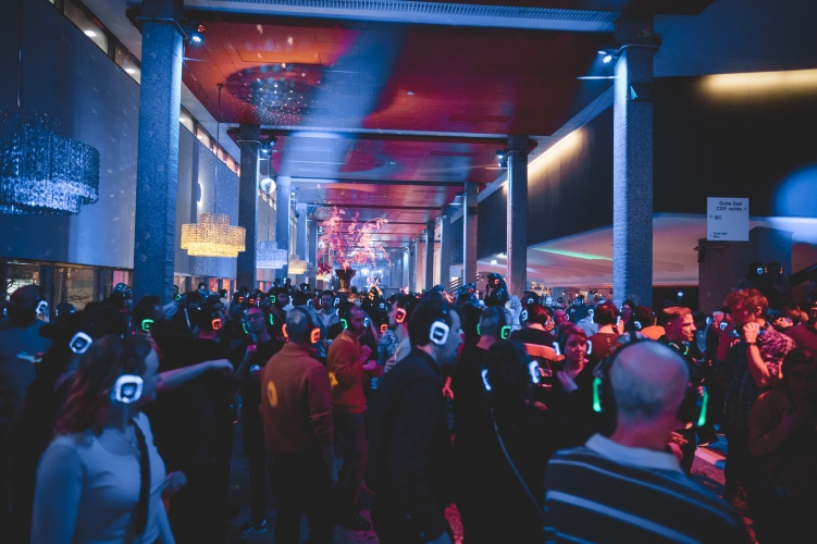 De foto toont de Grote Zaal Foyer van de Doelen in Rotterdam tijdens een evenement. De ruime foyer is gevuld met bezoekers die een silent disco beleven en een koptelefoon dragen. De ruimte heeft hoge kolommen, een rood plafond, marmeren vloeren en grote kroonluchters. Gekleurde verlichting zorgt voor een feestelijke sfeer. De foyer is geschikt voor ontvangsten, borrels en grootschalige events.