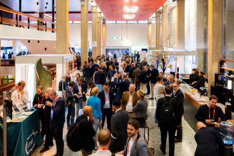 De foto toont de Grote Zaal Foyer van de Doelen in Rotterdam tijdens een congres. In de foyer staan moderne expositiestands met presentatieschermen en informatiebalies, waar bezoekers met elkaar in gesprek zijn. Boven de stands hangt een grote kristallen kroonluchter. De ruimte heeft hoge kolommen, lichte wanden en een ruime opzet, geschikt voor exposities, netwerkbijeenkomsten en ontvangsten.