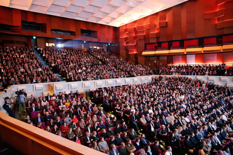 De foto toont de Grote Zaal van de Doelen in Rotterdam tijdens een uitverkocht evenement. De zaal is volledig gevuld met bezoekers die in vaste rijen rode stoelen zitten, verdeeld over meerdere niveaus en balkons. De wanden zijn bekleed met warme houten panelen en sfeervolle verlichting. De Grote Zaal heeft een imposante uitstraling en is geschikt voor grootschalige congressen, conferenties en culturele evenementen.