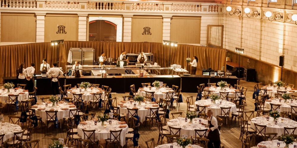 Sfeervol ingerichte evenementenhal met ronde gedekte tafels, witte tafellakens en stoelen, geschikt voor een groot diner, bruiloft of zakelijk event, inclusief professionele tafelaankleding en meubilair van Accuraat Verhuur.
