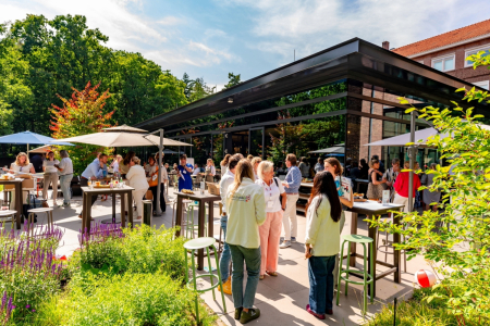 Het terras van restaurant KonneKt bij Conferentiehotel Kontakt der Kontinenten, waar mensen staand met elkaar in gesprek zijn aan hoge tafels, omgeven door groen, parasols en het moderne restaurantgebouw in een bosrijke omgeving.