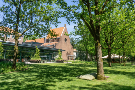 De bostuin en het terras van Conferentiehotel Kontakt der Kontinenten, met tafels en stoelen bij restaurant KonneKt, een zitzak op het gras op de voorgrond en een evenemententent op de achtergrond, omgeven door bomen in een bosrijke omgeving.