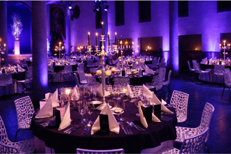 De foto toont een grote evenementenruimte in een kapel met een hoog, gebogen plafond dat rijk is beschilderd met klassieke en artistieke motieven. De ruimte is uitgelicht in blauwe tinten en voorzien van professionele verlichting en geluid. De diner opstelling en het hoge plafond geven een indrukwekkende, chique sfeer, geschikt voor diners, borrels en grootschalige evenementen.