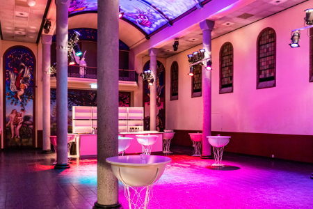 De foto toont een ruime evenementen locatie in een voormalige kapel met hoge zuilen, een beschilderd gewelfplafond en artistieke muurschilderingen. De zaal is verlicht met roze en paarse podiumverlichting. Er staan moderne, transparante statafels en achterin is een strak verlichte bar zichtbaar. De combinatie van historische architectuur en moderne inrichting maakt de locatie geschikt voor feesten, evenementen en culturele bijeenkomsten.