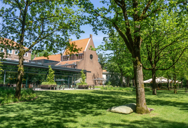 De bostuin en het terras van Conferentiehotel Kontakt der Kontinenten, met tafels en stoelen bij restaurant KonneKt, een zitzak op het gras op de voorgrond en een evenemententent op de achtergrond, omgeven door bomen in een bosrijke omgeving.