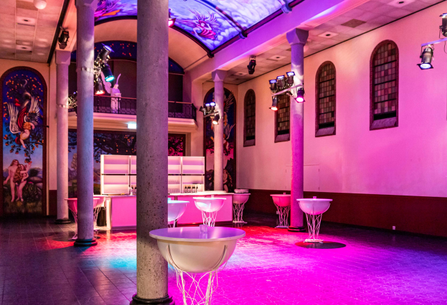 De foto toont een ruime evenementen locatie in een voormalige kapel met hoge zuilen, een beschilderd gewelfplafond en artistieke muurschilderingen. De zaal is verlicht met roze en paarse podiumverlichting. Er staan moderne, transparante statafels en achterin is een strak verlichte bar zichtbaar. De combinatie van historische architectuur en moderne inrichting maakt de locatie geschikt voor feesten, evenementen en culturele bijeenkomsten.